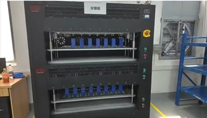 Inside a clean and modern battery manufacturing facility, rows of automated capacity grading cabinets (sorting machines) are testing lithium-ion battery cells. Numerous cylindrical or prismatic battery cells are neatly loaded into the testing slots of the equipment, with LED indicator lights showing the test status. A worker wearing anti-static clothing and a face mask may be seen monitoring the control screen or managing the loading and unloading process.