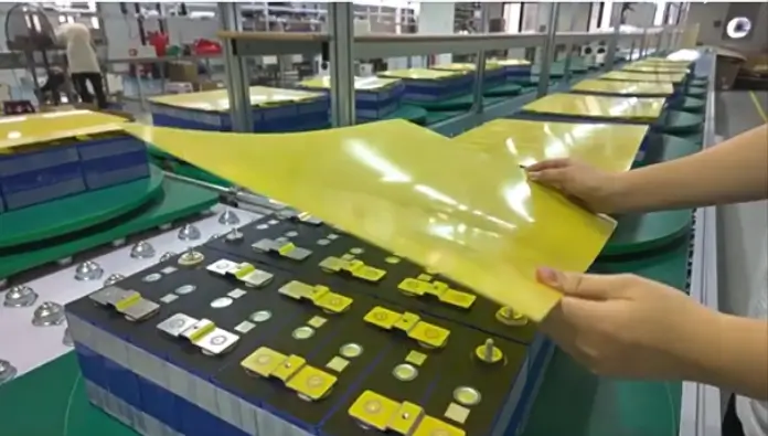 A worker on an electronics production line is carefully placing a thin, non-conductive insulation sheet (or insulating plate) onto a lithium-ion battery pack. The worker's hands are shown precisely aligning the insulation layer over the battery cells or busbars to prevent electrical short circuits, in a clean and well-lit workshop environment.